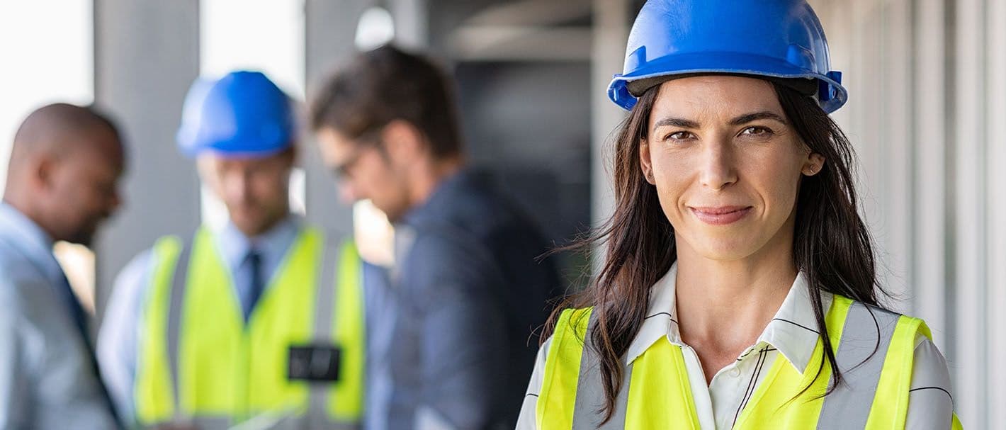 Portrait of a female architect standing on a construction site, smiling confidently. In the background, a group of men are looking at a plan. The focus is on the woman.